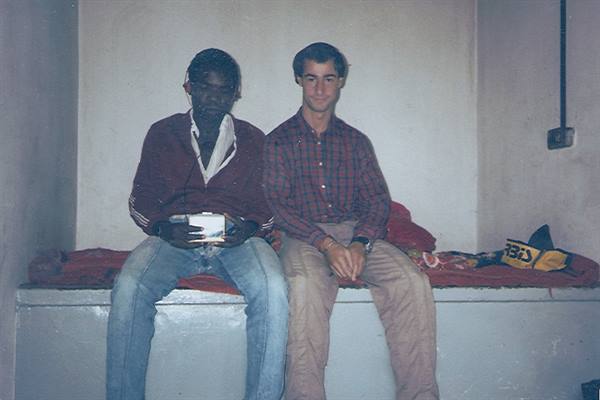 Two young men sitting on a bed in a bare room, one holding a photograph