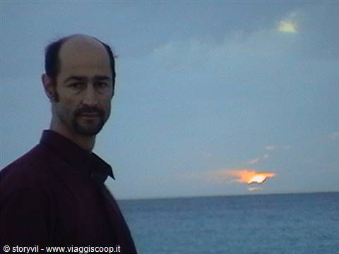 Man in dark shirt by the sea, orange glow behind clouds on the horizon
