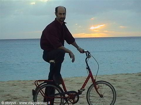 Man in maroon shirt riding a red folding bike on the beach under low sun