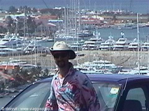 Man in straw hat by a car, marina full of yachts behind