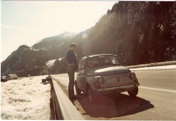 Man standing beside a parked Fiat 500 on a snowy roadside, forested mountains