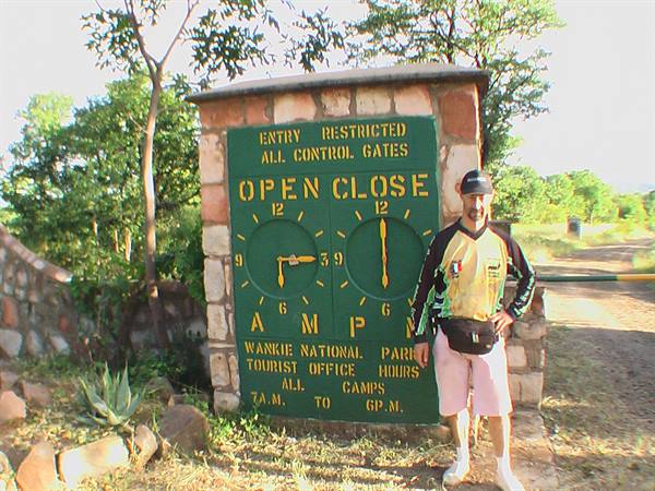 Man posing beside the entry times sign at Hwange National Park gate