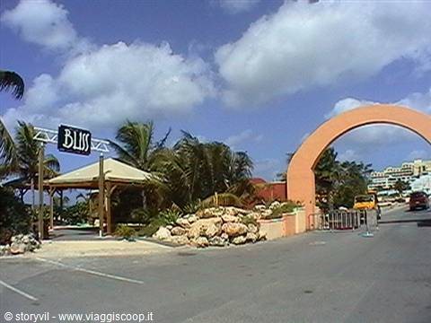 Salmon-pink arch and Bliss sign among palm trees and rocks, sunny tropical road