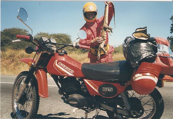 Motorcyclist in red helmet holding up a monitor lizard beside a Suzuki 185