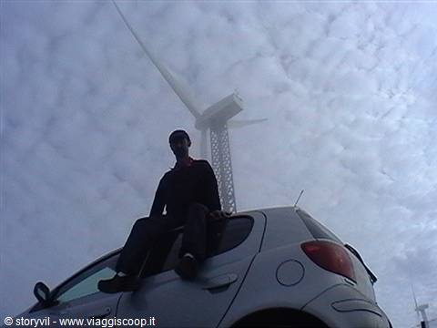 Man sitting on a gray car next to a huge wind turbine among clouds
