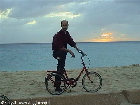 Man in red shirt standing beside red bicycle on the beach at sunset