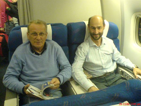 Two smiling men in blue airplane seats, one holding a magazine