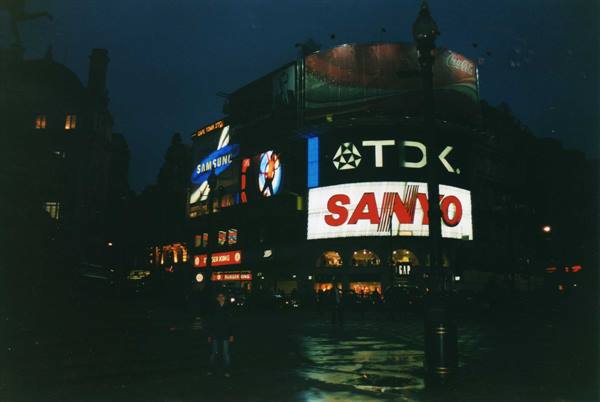 Piccadilly Circus at night: glowing TDK and Sanyo billboards on curved building, wet street
