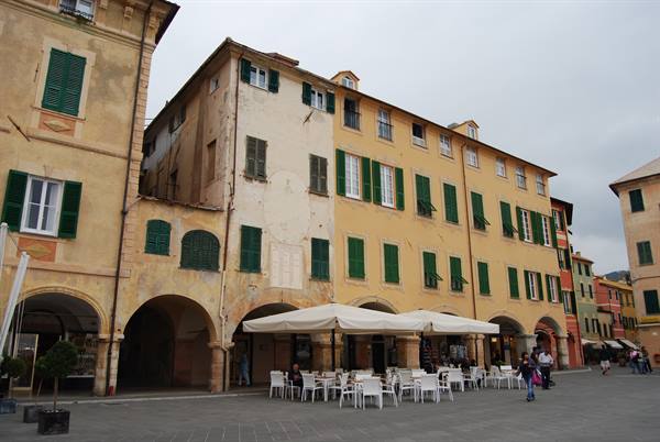 Ochre buildings with green shutters and arcades, white café tables under canopies