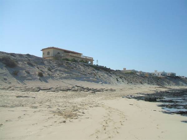 Ochre villa on sandy dune, empty beach with rocks under blue sky