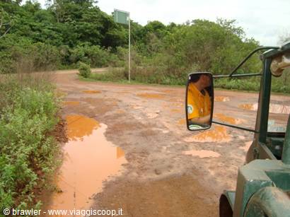Jeep side mirror with driver in yellow, muddy red road puddles in green forest