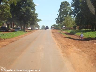 Red dirt road with cars passing, green trees and a child walking