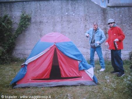 Two guys setting up a red and blue tent on grass by a concrete wall