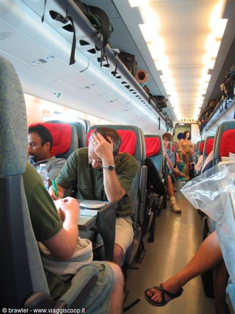 Passengers seated in red train seats, one writing under ceiling lights