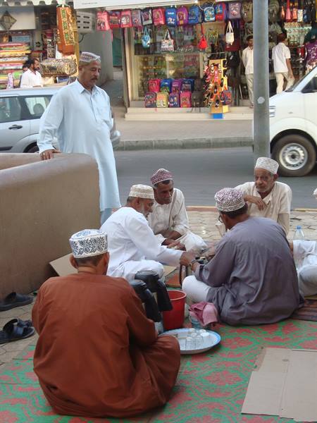 Omani men in embroidered caps sit on a mat by street shops