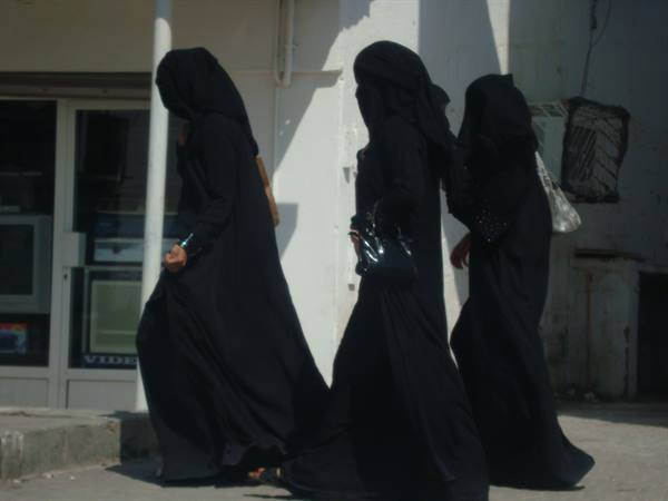 Three women in black abayas walk past a shop in bright sunlight