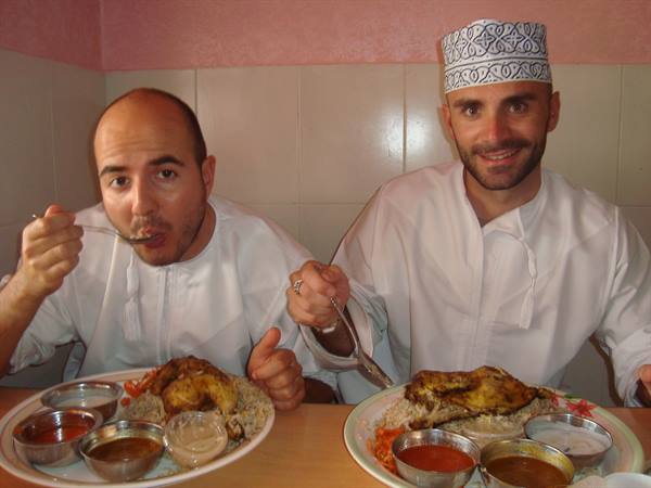 Two men in white garments with embroidered cap eating chicken and rice