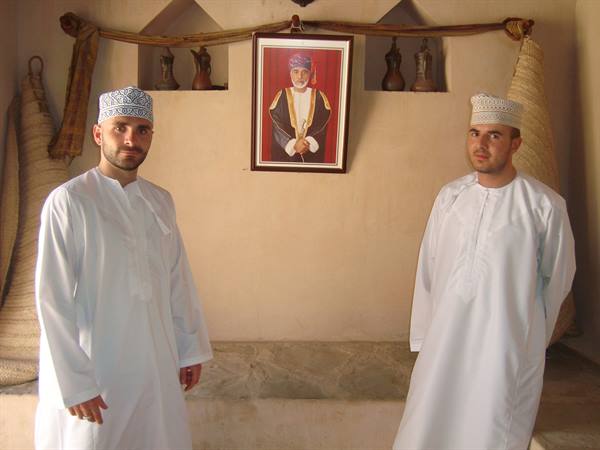 Two men in white dishdasha and embroidered caps beneath a portrait of Sultan Qaboos