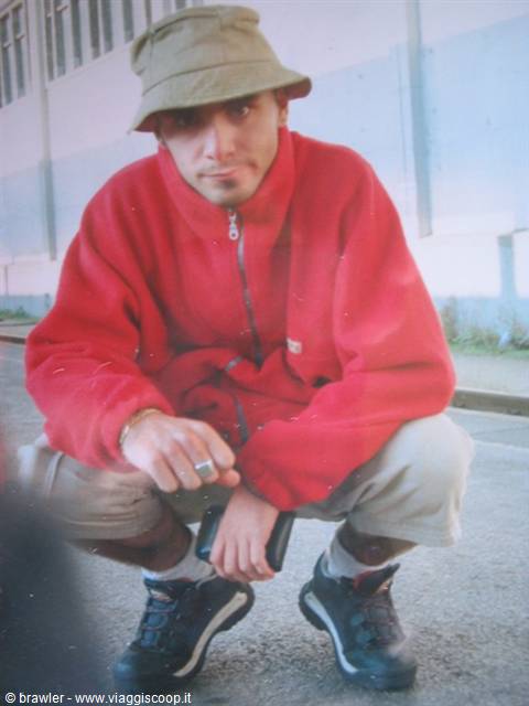 Man crouching in bucket hat and red fleece, in front of a pale building