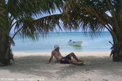 Man resting under leaning palms on white sand, boat anchored in turquoise sea