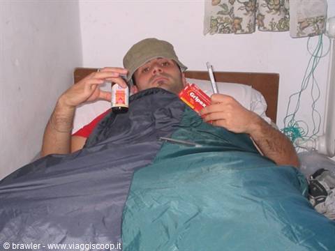 Man in cap in bed inside sleeping bag, holding beer and Gupp snacks
