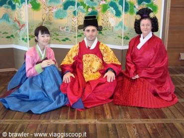 Three people in traditional hanbok sitting on wooden floor before a painted folding screen