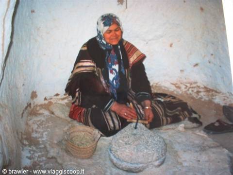 Woman in headscarf grinding grain on stone inside rustic white room