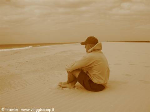 Man in cap sitting on sand gazing at the sea under cloudy sky