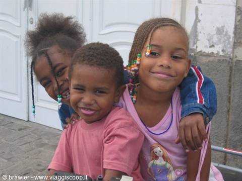 Three smiling children hugging together near a white door, cobblestone alley