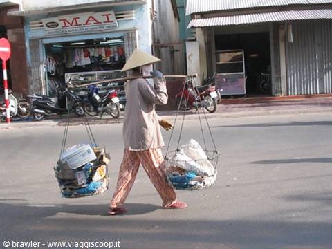 Woman in conical straw hat carrying two baskets of cardboard along motorbikes