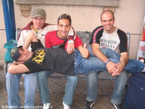 Four friends laughing on a metal bench, one lying across with thumbs up