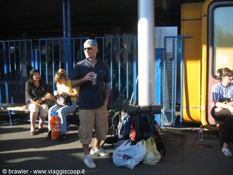 Travelers with backpacks and bags wait beside a yellow train at the station