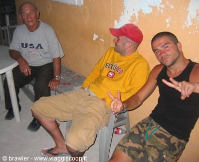Three men sitting in the shade against a weathered yellow wall, one flashing a peace sign