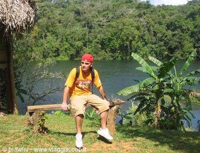 Tourist in red cap sitting on wooden bench in front of green jungle lake