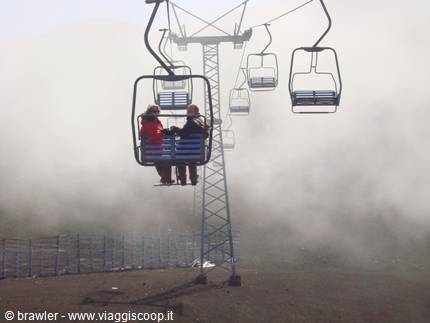 Two people on a blue chairlift rising through thick fog on Mount Etna