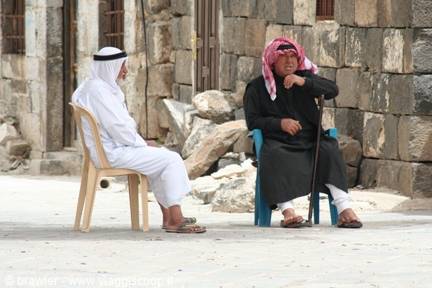 Two men in keffiyehs sitting on plastic chairs in front of an old stone wall
