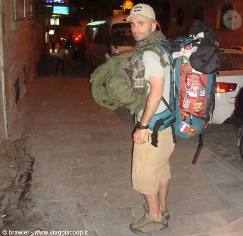 Backpacker with red backpack and green duffel walks in a narrow street at night