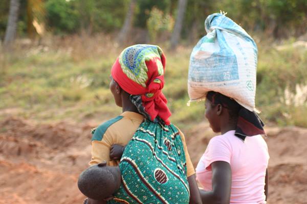 Two women walk on a red dirt road, one balancing a sack on her head