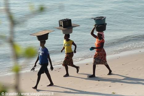Three women walk along the beach balancing plastic containers on their heads