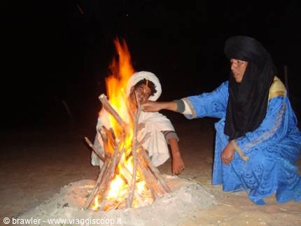 Woman in blue dress lights a wood fire beside a man in the desert at night