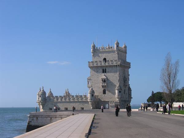 Tourists stroll beside the stone Belém Tower under a clear blue sky