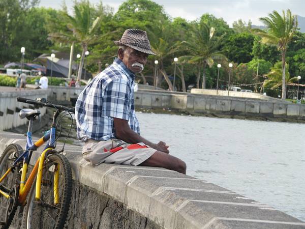 Elderly man in hat and plaid shirt sitting on stone pier next to yellow bike