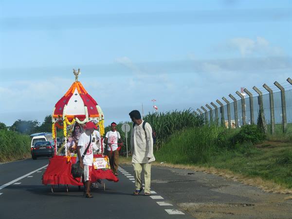 Pilgrims pull a red decorated cart with garlands along a roadside by cane fields