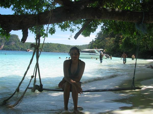 Woman in black swimsuit on wooden rope swing over turquoise water, boat behind
