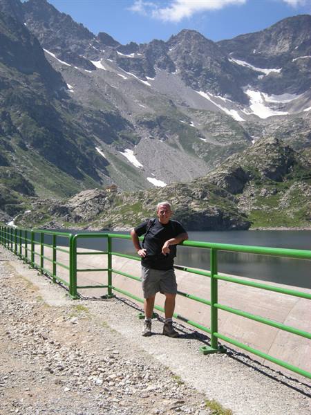 Man leaning on green dam railing, small lake and snowy peaks behind