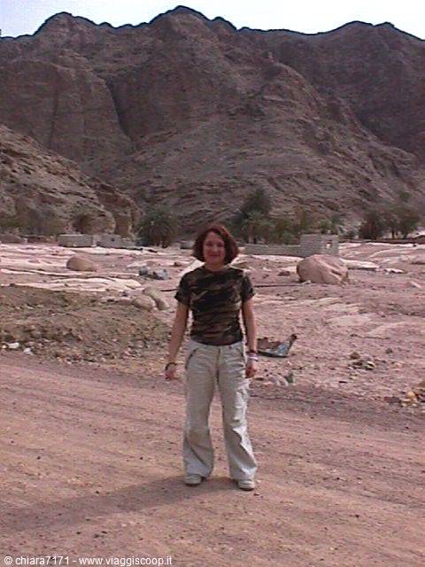 Woman in military shirt and white jeans on sandy road, rocky mountains behind