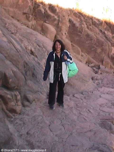 Woman in blue jacket with green bag standing on red stone path among desert rocks