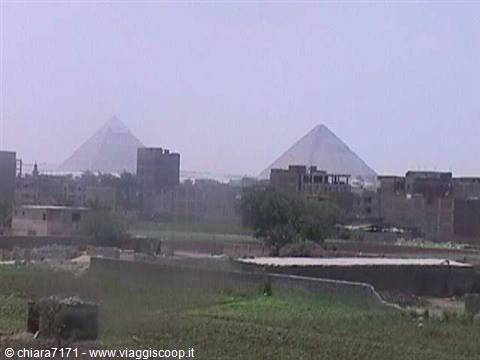 Pyramids of Giza rising above brick buildings, hazy sky