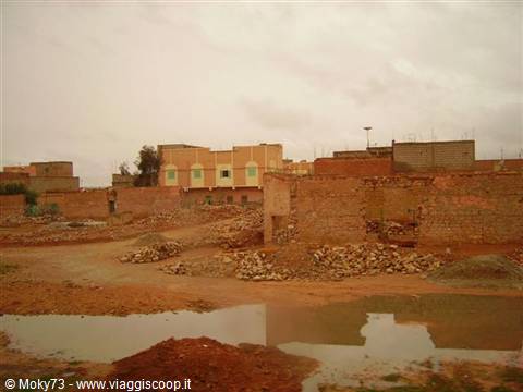 Mud-brick houses amid rubble and puddles under a grey rainy sky