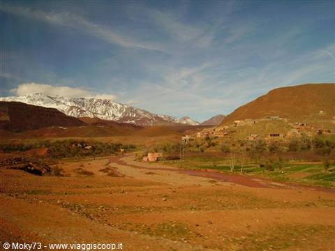 Mud-brick village among ochre dunes, snow-capped Atlas mountains in the background